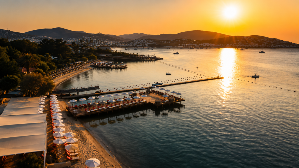 Luxury beach club in Bodrum with a private pier, white umbrellas, sun loungers, crystal-clear sea, and a stunning sunset over the Aegean coastline.