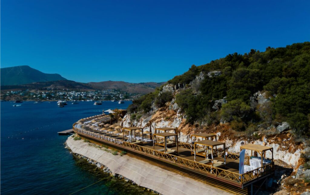 Ministry of Culture and Tourism Türkbükü Public Beach featuring crystal-clear turquoise waters, a clean sandy shoreline, sun loungers, and a relaxing public beach atmosphere in the heart of Bodrum.