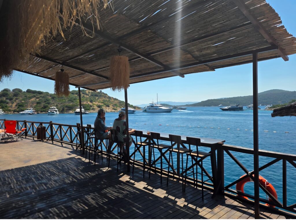 Türkbükü Public Beach featuring crystal-clear turquoise waters, a clean sandy beach, comfortable sun loungers, and a relaxing seaside atmosphere in the heart of Bodrum.