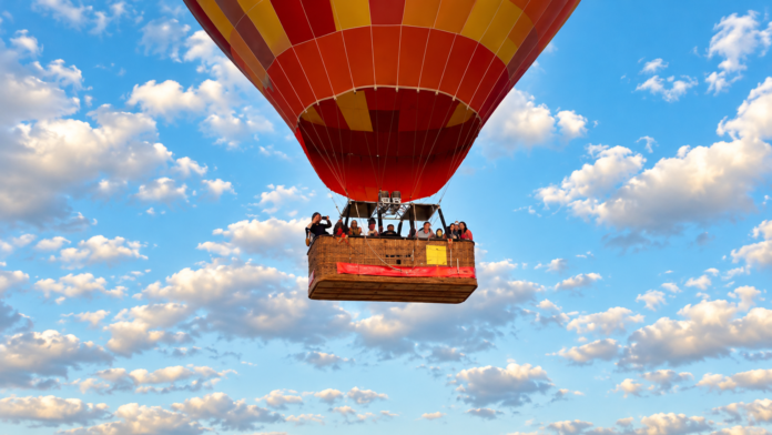 Red and orange hot air balloon carrying passengers against a blue sky with scattered clouds.